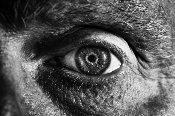 Black and white toned image depicting an extreme close up macro view of the eye of a senior man in his 70s. The man has blue eyes and they are a little bit bloodshot with the capillaries visible.