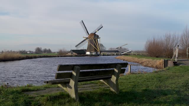 Traditional dutch windmill in the village of Baambrugge rotating, empty bench and Angstel River in the foreground