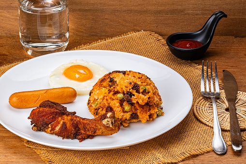 American fried rice with fried drumstick, fried egg and sausage in white ceramic plate with ketchup in black cup, a glass of wter, metal fork and knife on sack cloth.