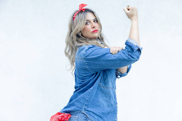 With a determined gaze and a powerful fist raised, a woman in a denim outfit stands as a modern symbol of the strength and empowerment that defines today's femininity.