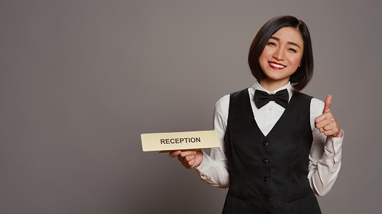Hotel concierge holding sign to indicate direction for reception desk, presenting indicator to help guests and offer assistance in studio. Employee with uniform and bow uses wall pointer. Camera B.