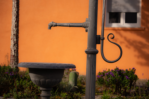 Details of a simple standing fountain in a small flower island. Detail of the water dispenser. Can be seen: Pump arm and round water trough in front of house facade. Close-up in fall.
