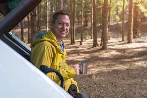 A happy man with a mug of tea or coffee sits in the open trunk of a car in the forest