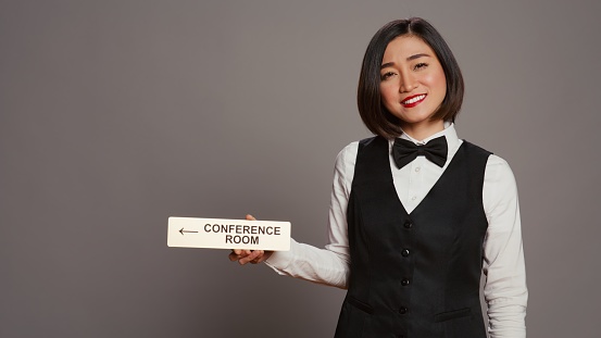 Asian receptionist pointing at wall indicator for conference room, using pointer to help customers find all amenities and facilities. Woman employee holding sign to indicate directions. Camera B.