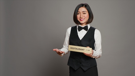 Asian hotel concierge holding restaurant sign to indicate direction, pointing towards dining area. Receptionist assisting clients to enjoy all amenities, stands over grey background. Camera A.