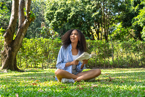 Happy young Asian woman spending her free time in weekend by sitting on grass in national green park while reading a book to relax and feeling happy with a fresh air. Woman in casual cloth enjoys moment in nature with love and realizes to protect nature.