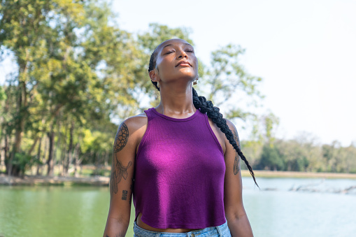 Beautiful young adult African American woman in sportwear closing her eyes and taking a deep breath with fresh air while sitting on wooden log near lake in green park. She is feeling happy and passionate commitment to protect nature.