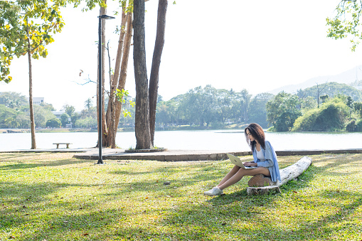 Young Asian woman in casual blue cloth sitting in park among beautiful green trees on the wooden timber and working on laptop to communicate with her team online in fresh air and beautiful nature environment. Young Asian woman enjoys moment in nature with love and realizes to protect nature.