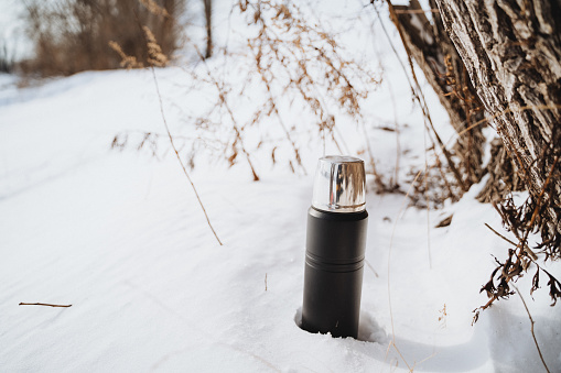 A black thermos with a silver lid is placed in the white snow near the tree trunk on a sunny winter day. The thermos is isolated on a white snow background, making it easy to see and identify.