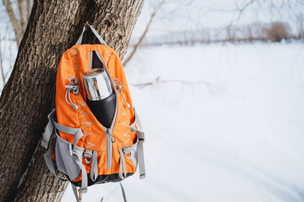 istock Orange backpack hanging on a tree in a snowy forest with a silver thermos bottle inside. 2007866325