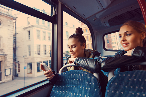 Female friends sitting on the bus, one of them using smart phone.