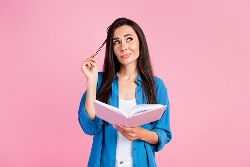 Photo of clever confused girl with straight hair dressed blue shirt hold pencil on head look empty space isolated on pink color background.