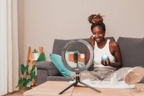 istock smiling african american Beauty Blogger Demonstrating Moisturizer Application sitting on sofa During online video skincare tutorial using smartphone with ring light 2007900972