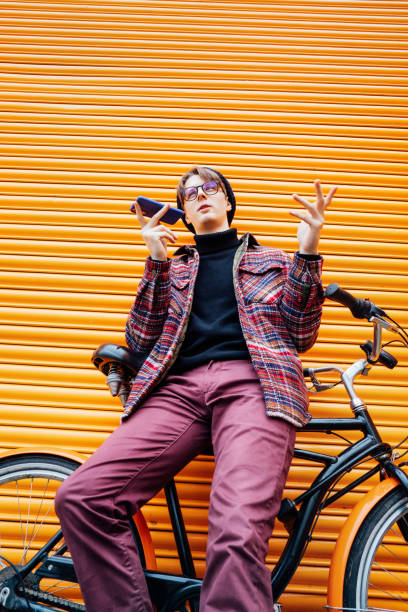 Young hipster man recording voice message on phone while sitting on cruiser bicycle near orange wall. Student using 5g network. Eco friendly transport for carbon footprint. Sustainable lifestyle.