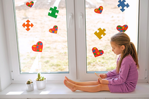 A sad lonely girl is sitting on the windowsill with a postcard in her hands. The windows of the room are decorated with hearts and colored puzzles for World Awareness Day about autism.