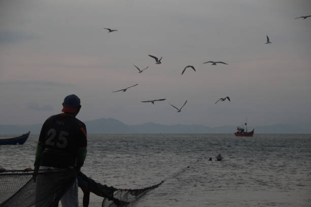 Banda Aceh, 12/13/2019, Fishermen are repairing their fishing nets on the beach at sunset. In the reddish-orange glow of twilight, they diligently clean the nets from the day's catch. A peaceful atmosphere is created amidst the shimmering sea, while the sky welcomes the night with captivating warmth