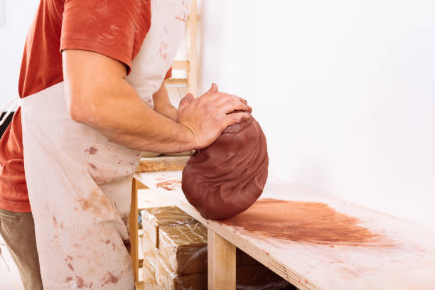 Crop of urecognizable, potter wearing apron on top of casual clothes and kneading soft clay on wooden board in workshop