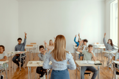 Multi-ethnic group of school children raising their arms to answer teacher's question on a class in the classroom. Childhood, education and people concept