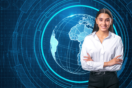 Happy young european businesswoman with folded arms and holographic screen with digital globe interface and business chart on blurry blue background. HUD, international and future monitor concept