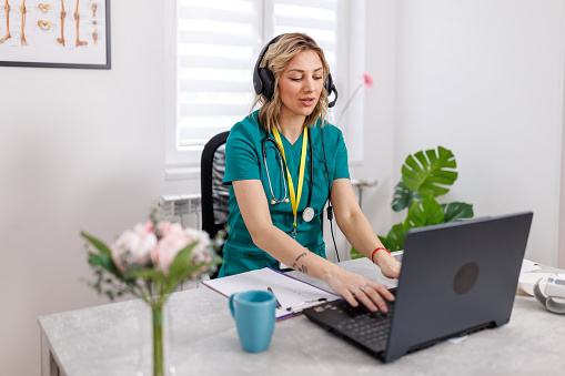 Utilizing headphones and her laptop, a female doctor engages in virtual consultations, her attention undivided as she listens and advises, marking a new era of patient interaction