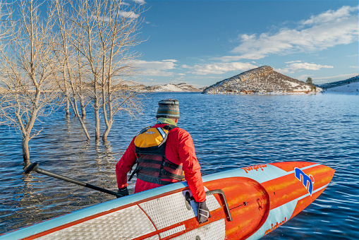 Fort Collins, CO, USA - February 15, 2020: Male paddler wearing a drysuit and life jacket is launching a long racing stand up paddleboard on a mountain lake in Colorado - Horsetooth Reservoir in winter conditions.