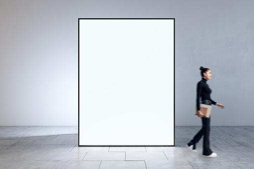Businesswoman walking in interior with modern blank big white mock up banner frame. Museums and exhibition concept