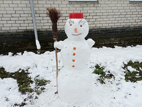 A snowman made of fresh white snow with a carrot nose and red pelvis on his head and holding a broom in his hands. Winter entertainment and snow sculptures.