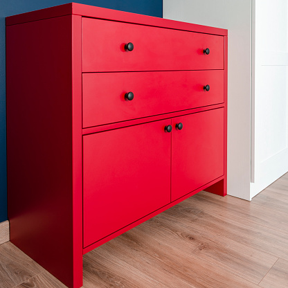 Red chest of drawers in a Scandinavian interior. A red wooden cabinet with black handles stands in an apartment against a blue wall next to a white wardrobe.