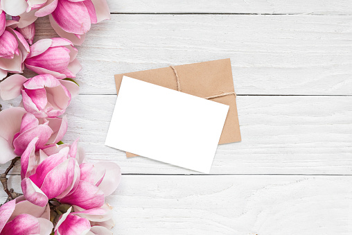 Blank greeting card with spring pink magnolia flowers on white wooden background. Mock up. Flat lay. Top view. Womens day, Mothers day, Wedding or anniversary invitation.