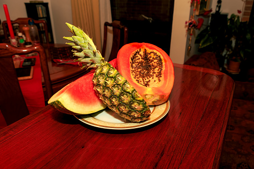 Half a ripe pineapple and papaya on a plate on a shiny rosewood table. To one side, there is a segment of watermelon. They are ready to eat.