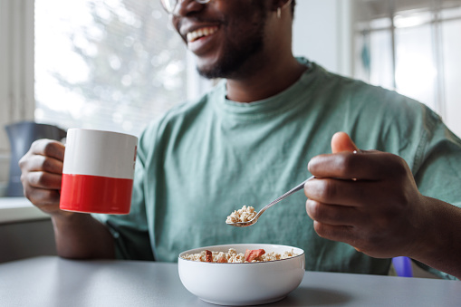 Young African American man having a breakfast. He is holding a cup of coffee and eating oatmeal