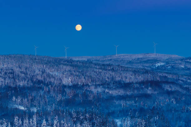 A full moon over a winter forest landscape with wind turbines on the horizon on an early winter morning. Dalarna region, Sweden.