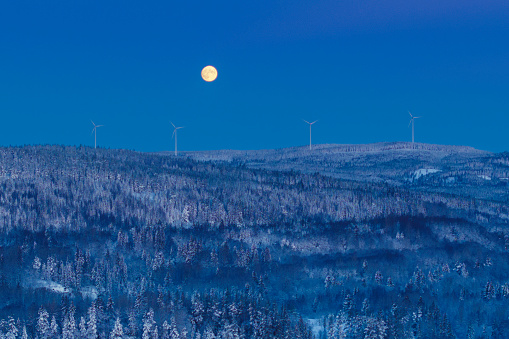 A full moon over a winter forest landscape with wind turbines on the horizon on an early winter morning. Dalarna region, Sweden.
