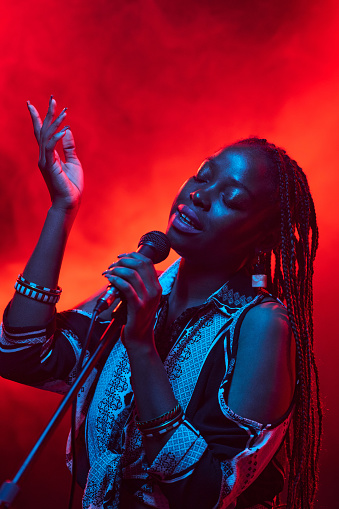 Vertical portrait of young Black woman singing into microphone performing on stage with smoke in background