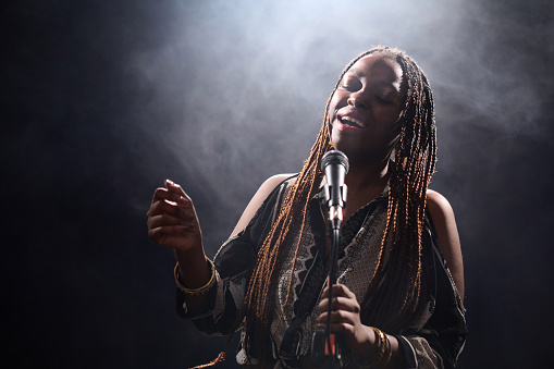 Waist up portrait of talented Black woman singing into microphone on stage with smoky background copy space