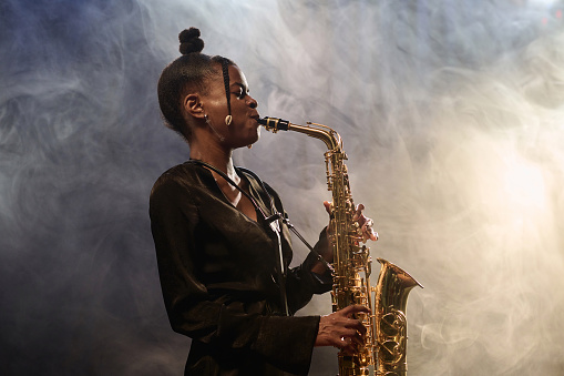 Side view portrait of talented African American woman playing saxophone in jazz music club with smoke effect copy space