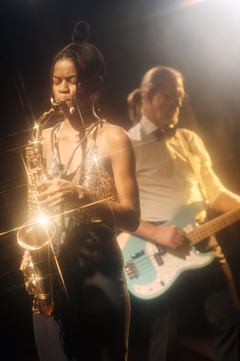 Vertical portrait of African American woman playing saxophone performing with jazz band on stage gold light accents