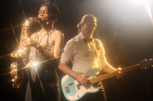 Portrait of young African American woman playing saxophone performing with jazz band on stage in hazy nightclub copy space
