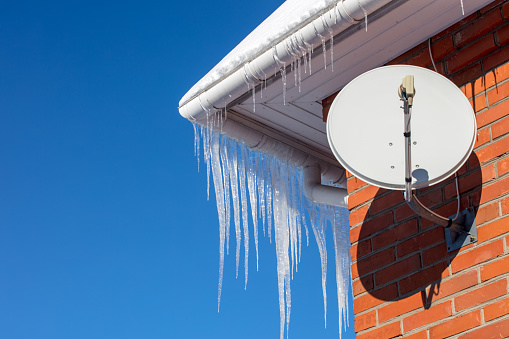Close-up of shiny icicles on a roof gutter of a brick house with a satellite dish. Space for copy on a blue sky.