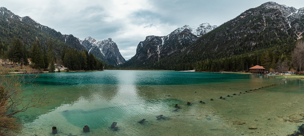 Panoramic view of turquoise water alpine lake Lago di Dobbiaco with restaurant in Dolomiti mountains, Cortina dAmpezzo, Italy on moody spring day. Lake Toblacher See in the forest in Dolomites in Italian Alps.