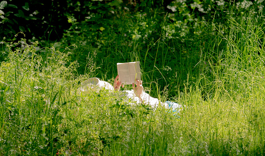 Cheerful caucasian student guy reading book outdoors, preparing for lectures while lying on lawn in university campus or park, free space