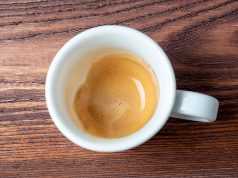 A white porcelain cup with coffee in it, on a wooden background. The foam is brown on the sides of the cup. A flavorful, invigorating breakfast drink. Top view, flat lay
