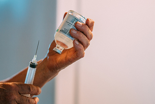 Close-up shot of Caucasian veterinarian woman getting ready to draw clear liquid medication from a glass vaccine vial container with a large hypodermic syringe indoors at an animal hospital