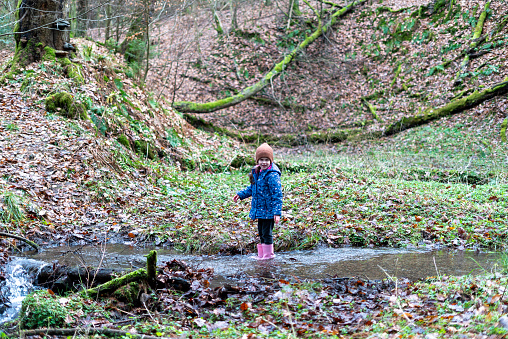 Little girl in a raincoat and rubber boots standing in a stream in the forest