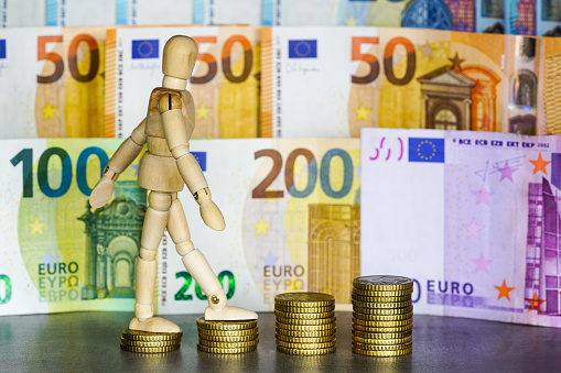 A wooden human figure climbs up stacks of coins of increasing height, euro banknotes background, concept of financial success