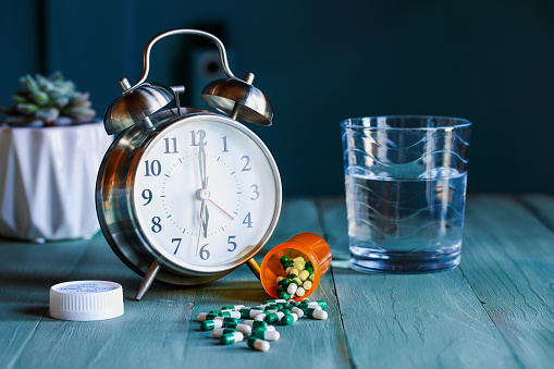 Open bottle of  prescription pills by an alarm clock and a glass of water on wooden night stand table. Selective focus with blurred background.