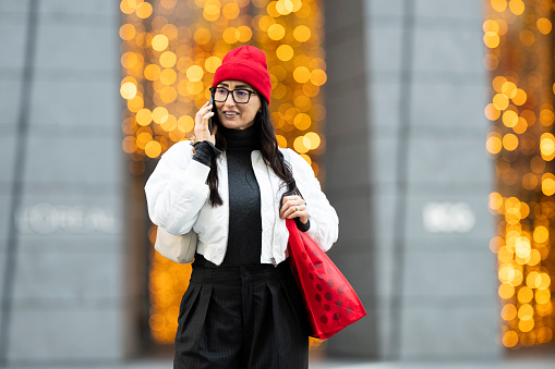 A beautiful woman wearing red hat, eyeglasses, white bomber jacket, black clothes and a red bag seen talking on the phone while walking at the Hudson Yards in New York City.