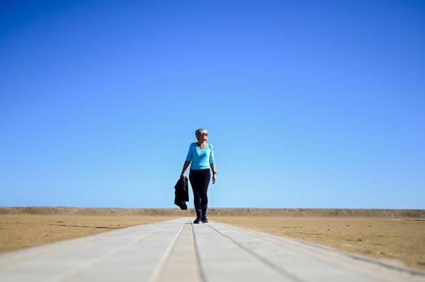 vue en contre-plongée d’une femme mûre marchant sur une plage de sable devant un ciel clair. - senior adult outdoors wellbeing sky photos et images de collection