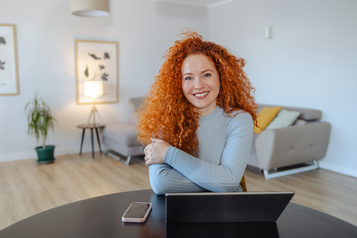Photo of young woman with red hair sitting at the table in the living room and using laptop. She is smiling and looking at the camera