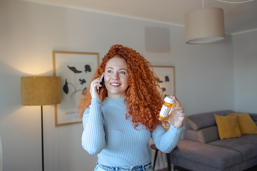 Photo of young woman talking on the phone at home and holding bottle of medicines